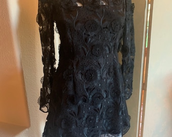 Pode incluir: Um vestido preto de renda floral de manga comprida, com decote barco e bainha festonada.