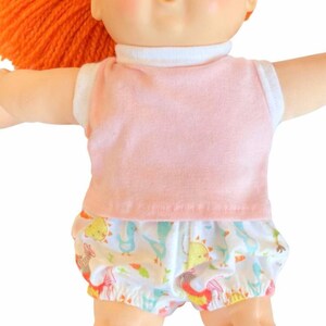 May include: A doll with bright orange yarn hair, green eyes, and a small open mouth. The doll wears a light pink sleeveless top with a white collar and white shorts featuring a colorful pattern of small cartoon animals and shapes.