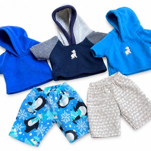 May include: Three short-sleeved hooded sweatshirts in blue, navy with grey, and bright blue, each featuring a small white bear graphic. Two pairs of shorts are included: one with a blue penguin and snowflake print, and one with a grey patterned design.