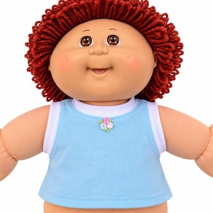 May include: A doll with a round face, brown eyes, and a wide smile, featuring thick, curly dark red yarn hair. The doll wears a light blue sleeveless top with a white trim around the neck and armholes, accented by a small floral embroidery at the center.