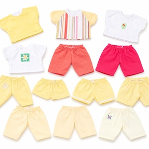 Peut inclure: Une collection de vêtements pour poupée comprenant trois hauts à manches courtes en jaune, blanc avec une fleur, et blanc à rayures, ainsi que neuf shorts dans diverses nuances de jaune, corail, rose et crème avec un papillon.