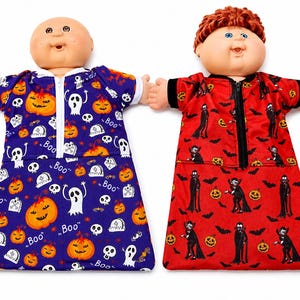 May include: Two dolls wearing Halloween-themed sleep sacks. The left doll wears a purple sleep sack with orange pumpkins, white ghosts, and the text "Boo". The right doll wears a red sleep sack with black vampire figures, bats, and orange pumpkins.