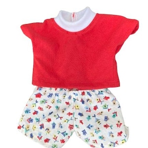 May include: A red short-sleeved top with a white collar and matching shorts. The shorts are white with a colourful pattern of birdhouses and other shapes. This outfit is suitable for a doll.