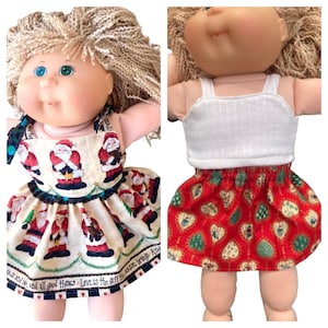 May include: A doll with curly blonde hair wearing two different outfits. The first is a cream-colored dress with a Santa Claus print and text along the hem. The second is a white tank top paired with a red skirt featuring a heart and tree pattern.