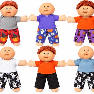 May include: Six dolls with light skin tones, round faces, and blue eyes. Some have red hair, others are bald. They wear various short-sleeved shirts in gray, purple, orange, white, or blue, paired with patterned pants featuring pumpkins, ghosts, or solid black.