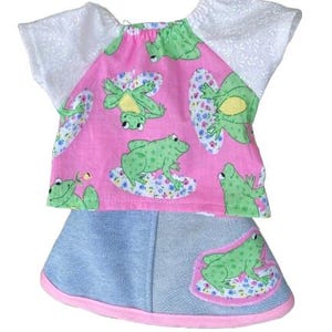 May include: A two-piece outfit consisting of a short-sleeved top and a skirt. The top features a pink fabric with a green frog pattern and white sleeves. The blue denim skirt includes a pink hem and an appliqué of a green frog on a floral patch.