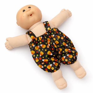 May include: A bald doll with a light skin tone wearing black dungarees featuring a repeating pattern of orange, yellow, and white candy corn shapes interspersed with small red and yellow dots. The doll has an open mouth and outstretched arms.