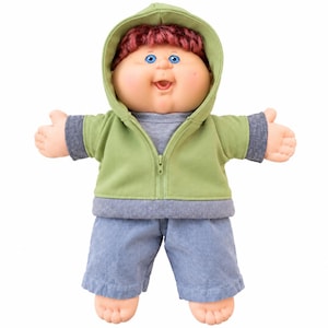 May include: A doll with a round face, blue eyes, and reddish-brown hair, wearing a light green hooded zip-up jacket over a gray shirt, paired with blue shorts. The doll has a light skin tone, an open mouth, and arms extended outward.
