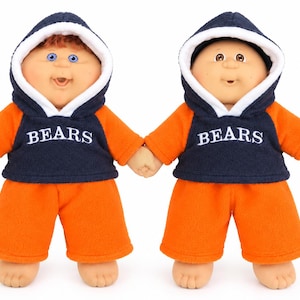 May include: Two dolls with light skin tones wearing matching navy blue and orange hooded sweatshirts with the word BEARS printed in white across the chest, paired with bright orange shorts. One doll has reddish-brown hair and blue eyes.