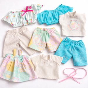 May include: A selection of doll clothing in pastel shades. The set includes shorts, trousers, tops, and a skirt. Some items display floral and geometric patterns, while others are plain. A pink ribbon is also included.