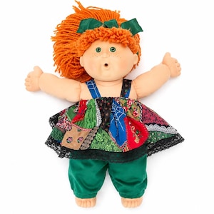 May include: A doll with bright orange yarn hair styled with a dark green bow, featuring green eyes and freckles. The doll wears a patchwork dress with various floral and geometric patterns in red, blue, and green, finished with black lace trim.