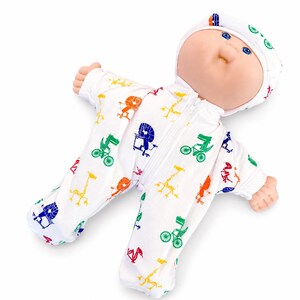 May include: A baby doll in a white onesie patterned with bicycles, giraffes, and other shapes in various colours. The doll has a matching white cap and a front zipper. The doll's face features blue eyes and a neutral expression.