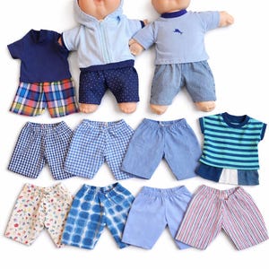 May include: Two dolls with round faces and light skin tones, one wearing a light blue hooded jacket and dark blue polka dot shorts, the other in a light blue t-shirt and denim shorts, accompanied by nine pairs of assorted patterned shorts and shirts.