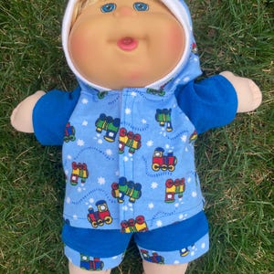 May include: A Cabbage Patch Kid doll wearing a blue hooded outfit with a train pattern. The doll has blonde hair and blue eyes.