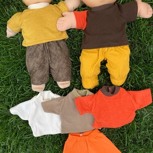 May include: Two Cabbage Patch Kids dolls with various outfits. One doll wears a yellow shirt and brown patterned pants. The other doll wears a brown shirt with orange sleeves and yellow pants. Additional shirts and pants in white, tan, and orange are also shown.
