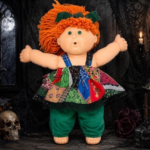 May include: A doll with bright orange yarn hair, a dark green hair bow, and green eyes. The doll wears a patchwork dress featuring various patterns including spiderwebs, floral prints, and geometric shapes, paired with solid dark green trousers.
