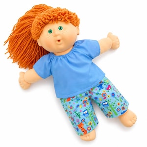 May include: A doll with bright orange yarn hair, green eyes, and freckles on its cheeks. The doll wears a solid light blue short-sleeved shirt and trousers featuring a colourful pattern of small owls, foxes, flowers, and green stems on a blue background.