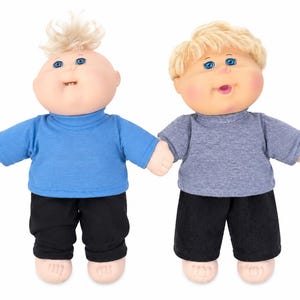 May include: Two dolls with round faces, blue eyes, and rosy cheeks. The left doll has spiky blonde hair, a blue mock-neck shirt, and black pants. The right doll has wavy blonde hair, a grey t-shirt, and black pants. Both dolls have bare feet.