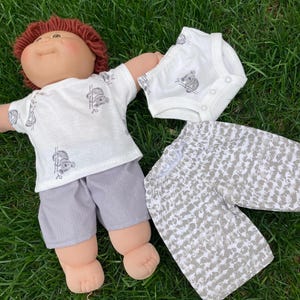 May include: A doll dressed in a white t-shirt and grey shorts, with a matching onesie and patterned trousers. The shirt and onesie feature a koala bear design. The doll is on green grass.