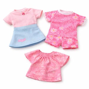 May include: Three sets of doll clothes. One set includes a pink t-shirt with a small flower and a light blue skirt with the word "Angel". Another set is a pink floral print top and shorts. The third is a pink floral top.