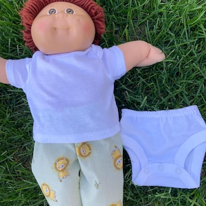 May include: A Cabbage Patch Kid doll wearing a white t-shirt, light green pants with yellow lion graphics, and white underwear. The doll has red hair and is lying on green grass.