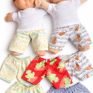 Puede incluir: Dos muñecas Cabbage Patch Kids con camisetas blancas y pantalones cortos estampados. Los pantalones cortos presentan diseños como notas musicales, ranas sobre fresas y patos. Los pantalones cortos son de varios colores, incluyendo rojo, azul y amarillo.