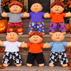 May include: Six Cabbage Patch Kids dolls with round faces and yarn hair, wearing various colored t-shirts and patterned shorts featuring Halloween motifs like ghosts, pumpkins, and vampires, arranged in two rows on a set of wooden steps.