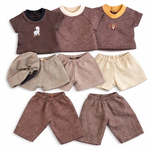 May include: A collection of doll clothing including three short-sleeved shirts in brown tones with crew necks, one featuring a small cat graphic and another an airplane. Includes three pairs of shorts, one hat, and two pairs of longer trousers.