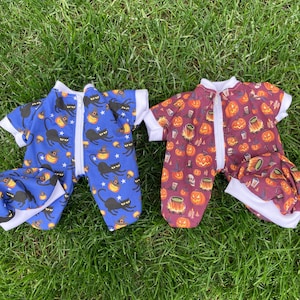 May include: Two Halloween baby sleepsuits on grass. One is blue with black cats, witch hats, and pumpkins. The other is maroon with jack-o'-lanterns, skulls, and potion bottles. Both have white trim and a full-length zip.