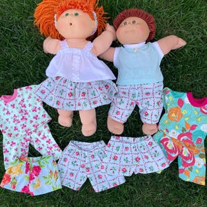 May include: Two vintage dolls with orange and brown hair, wearing matching outfits. The outfits include white tops, floral skirts, and shorts. Additional floral print pajamas and shorts are also displayed on a green grassy surface.