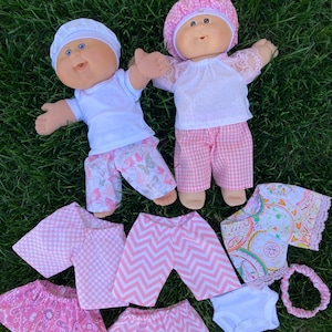 May include: Two Cabbage Patch Kids dolls wearing outfits, including white shirts, patterned pants, and hats. Additional doll clothes in pink and white patterns are arranged nearby, including skirts, shirts, and a headband. The dolls are on a green grassy surface.