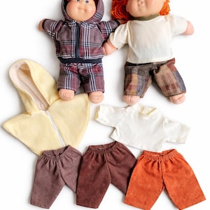 May include: Two vintage-style dolls with soft, round faces and colorful hair, dressed in plaid and solid-colored outfits. Additional clothing items include a yellow hooded jacket, white t-shirts, and various pairs of pants in shades of brown and orange.
