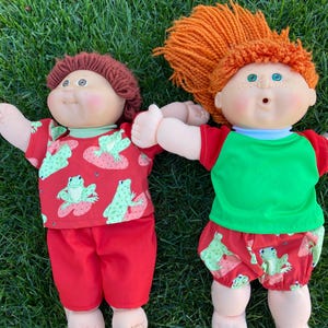 May include: Two Cabbage Patch Kids dolls lying on green grass. One doll has brown hair and wears a red outfit with a green frog and strawberry pattern. The other doll has orange hair and wears a red and green outfit with a frog and strawberry pattern.