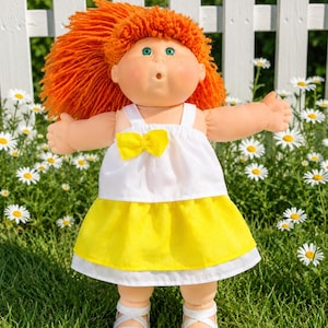 May include: A doll with bright orange yarn hair, green eyes, and freckles. The doll wears a white sleeveless top featuring a small yellow bow at the centre, a yellow skirt, and white strappy sandals. The doll has an open mouth expression.