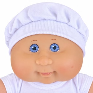 May include: A doll with a round, peach-toned face, bright blue eyes, and a small smiling mouth. The doll wears a white fabric hat with a ribbed brim and a matching white shirt. The doll has a soft, rounded appearance with visible dimples.