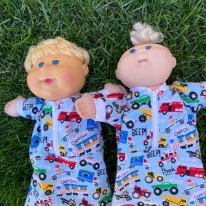 May include: Two Cabbage Patch Kids dolls wearing blue and white patterned pyjamas with a zip. The pyjamas have a print of various vehicles and the words "Choo Choo!" and "Beep!". The dolls are lying on green grass.