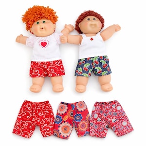 May include: Two dolls with yarn hair, one with orange hair and a white heart-print top, the other with brown hair and a white tank top. They wear patterned shorts. Three additional pairs of shorts with red, floral, and geometric prints are included.