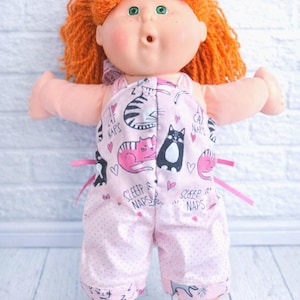 May include: A doll with bright orange yarn hair and green eyes wearing light pink overalls featuring a repeating pattern of cats, small hearts, and the text "I CAT NAPS" and "SLEEP NAPS". The doll has a rounded face, freckles, and an open mouth.