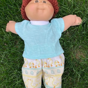 May include: A Cabbage Patch Kid doll with brown yarn hair, wearing a light blue t-shirt and patterned pants. The pants feature a repeating pattern of yellow ducks and green frogs on a white background. The doll is lying on green grass.