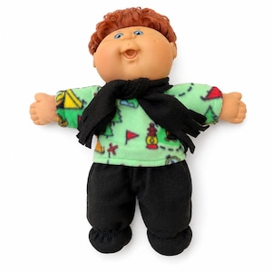 May include: A doll with curly reddish-brown hair and blue eyes, wearing a light green long-sleeve shirt featuring a colourful camping-themed print, a black fringed scarf around the neck, and solid black trousers with gathered cuffs at the ankles.