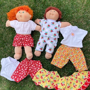 May include: Two Cabbage Patch Kids dolls with red and orange yarn hair, wearing white tops and patterned shorts. Additional outfits include red polka dot shorts, yellow cherry shorts, and orange floral shorts.