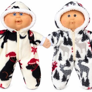 May include: Two dolls with round faces and blue or brown eyes, each wearing a hooded fleece sleeper with a front zipper. One sleeper features a black dog pattern with red Santa hats, while the other displays a grey deer and pine tree print.