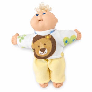 May include: A doll with a round, peach-toned face, blue eyes, and light blonde hair. The doll wears a white long-sleeved shirt featuring a brown lion graphic and green and blue patterns, paired with solid light yellow pants on a white background.