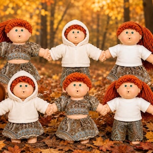 May include: Six dolls with orange yarn hair and green eyes arranged in two rows. The dolls wear various outfits including white hooded jackets, white t-shirts, brown paisley print crop tops, skirts, and shorts, all standing on a bed of autumn leaves.
