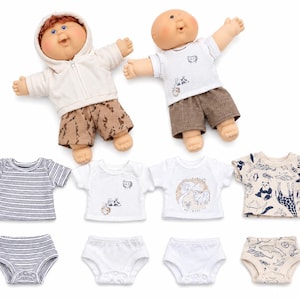 May include: Two dolls with round faces and yarn hair, one wearing a cream hoodie and brown camouflage shorts, the other in a white graphic t-shirt and brown shorts. Included are four additional t-shirts and four pairs of underwear with various patterns.