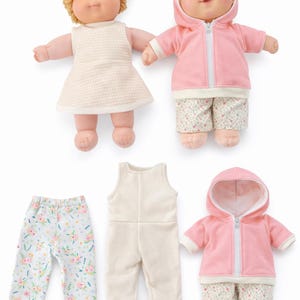 May include: Two dolls with blonde hair and blue eyes, one in a white dress, the other in a pink hooded jacket and floral trousers. Additional outfits include floral trousers, a cream jumpsuit, and a pink hooded jacket.