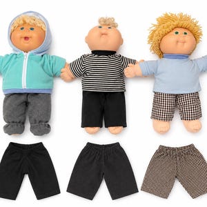 Peut inclure: Trois poupées aux cheveux en laine et aux traits doux. La première porte un sweat à capuche bleu sarcelle et un pantalon gris, la deuxième un t-shirt rayé noir et blanc et un short noir, la troisième un t-shirt bleu clair et un short à carreaux.