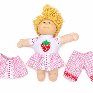 May include: A doll with yellow yarn hair and green eyes wearing a white top with a red strawberry graphic and a pink patterned skirt. Included are an additional matching pink patterned skirt and a pair of pink patterned shorts with lace trim.