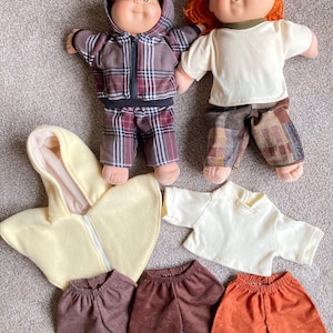 May include: Two Cabbage Patch Kids dolls with red and brown hair, wearing outfits. One doll is in a plaid jacket and pants, the other in a white shirt and patchwork pants. Additional doll clothes include a yellow hooded cape, white shirt, and three pairs of pants.
