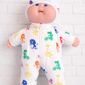 May include: A Cabbage Patch Kids doll wearing a white onesie and matching hat. The onesie features colorful illustrations of lions, giraffes, and alligators riding bicycles. The doll has blue eyes and a smiling expression.